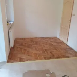 Wnętrze pokoju z ułożoną parkietową podłogą w jodełkę, widoczne narzędzia i kartony na nieukończonej części posadzki, surowa drewniana ościeżnica drzwiowa, białe ściany i kaloryfer.