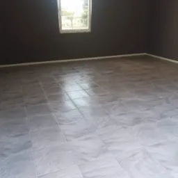 Pokój po remoncie z szarymi ścianami i nową, jasną podłogą z płytek imitujących marmur, widok na okno z zielenią na zewnątrz.