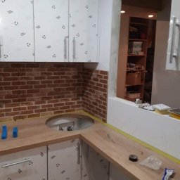 Ben-Bud - Montaż ceglanej ściany nad blatem kuchennym z wyciętym otworem na zlew, szafki górne w folii ochronnej, widoczne narzędzia i materiały na blacie.