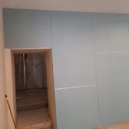 Ściana z płyt gipsowo-kartonowych w trakcie montażu, widoczne łączenia między płytami i otwór drzwiowy prowadzący do pomieszczenia ze stalową konstrukcją.