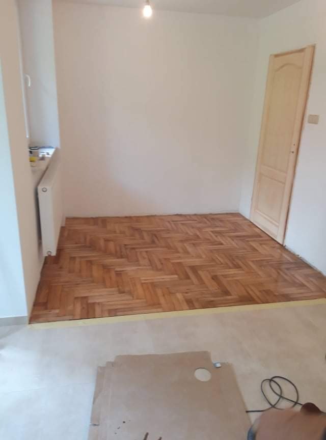 Wnętrze pokoju z ułożoną parkietową podłogą w jodełkę, widoczne narzędzia i kartony na nieukończonej części posadzki, surowa drewniana ościeżnica drzwiowa, białe ściany i kaloryfer.