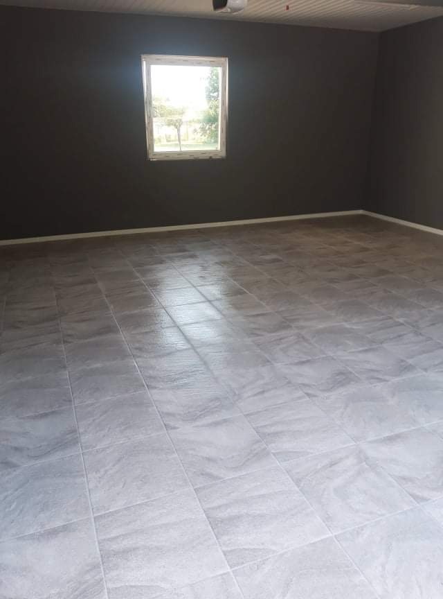 Pokój po remoncie z szarymi ścianami i nową, jasną podłogą z płytek imitujących marmur, widok na okno z zielenią na zewnątrz.