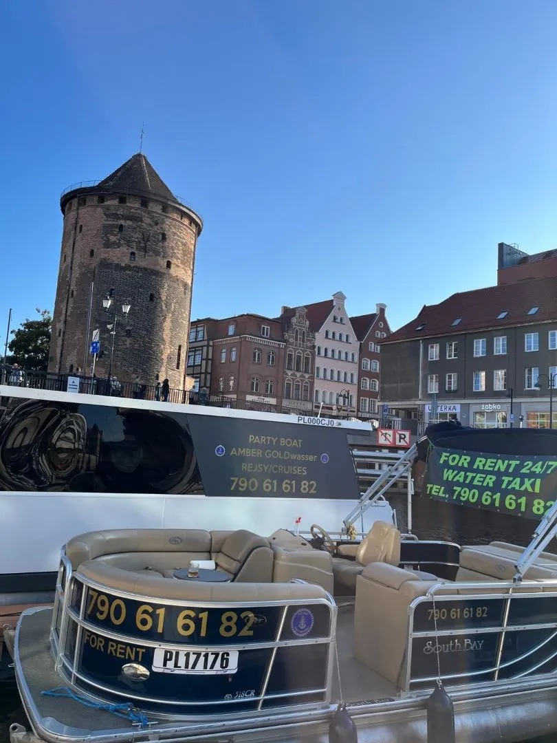Ponton turystyczny z numerem telefonu na wynajem, na tle starego miasta z wieżą, w słoneczny dzień.