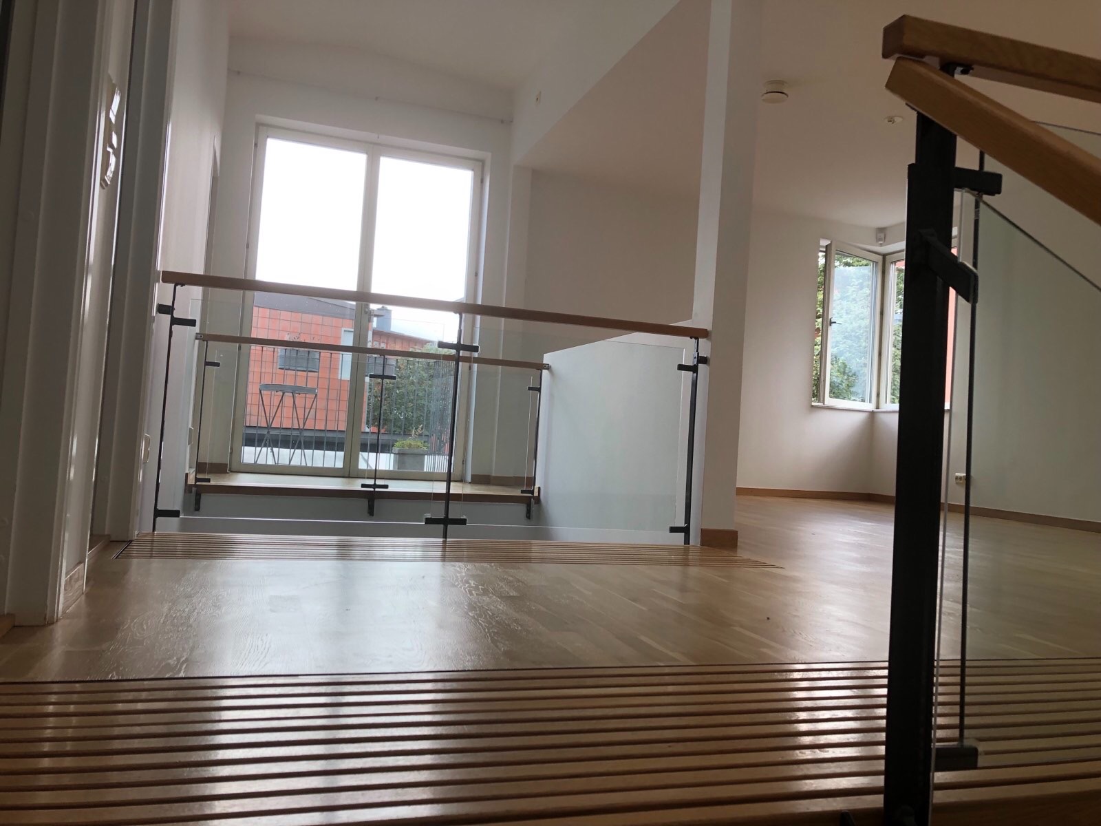 Jasne wnętrze domu z drewnianą podłogą i szklaną balustradą, widok z góry schodów na otwartą przestrzeń z oknami.