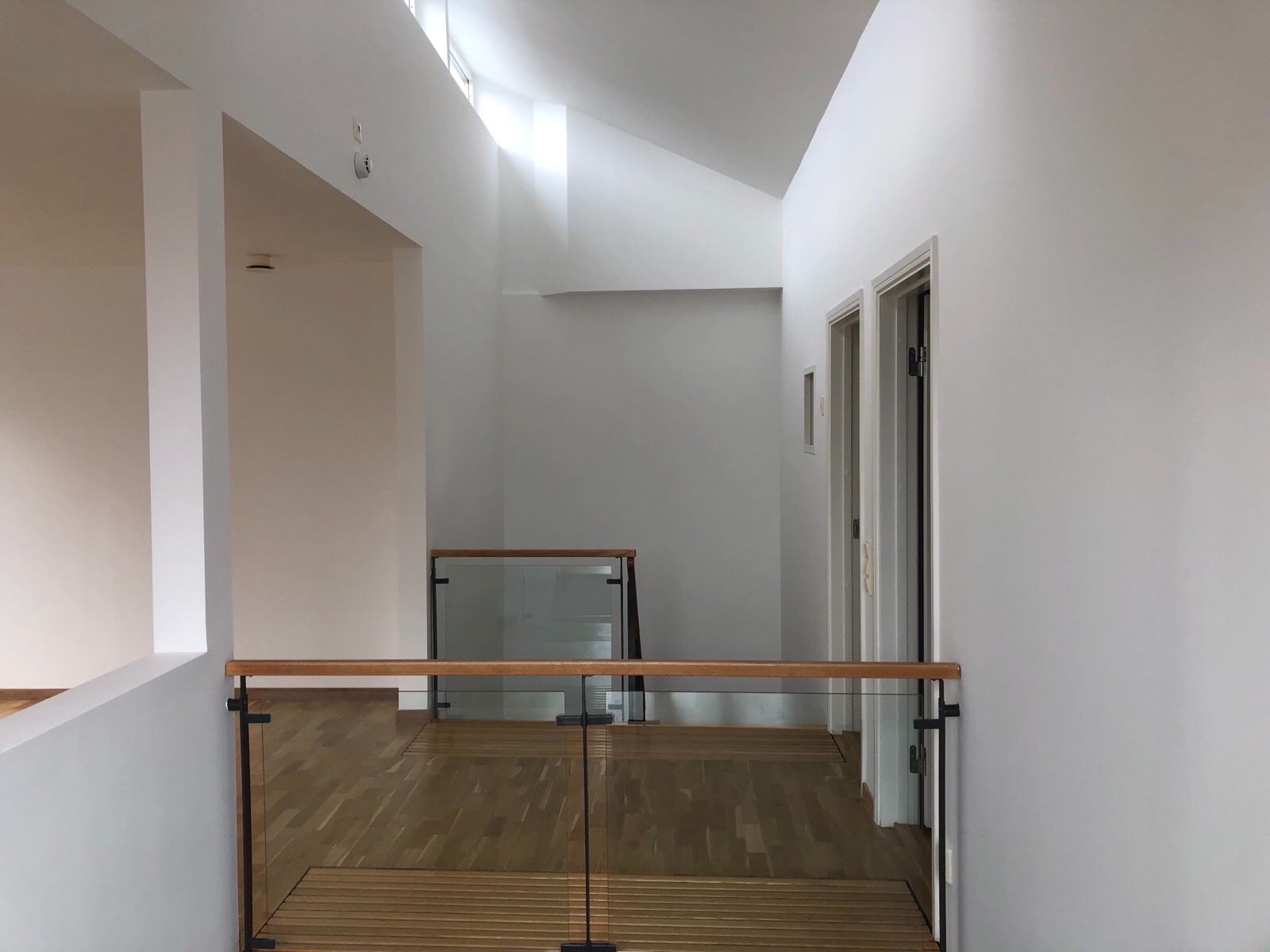 Wnętrze domu po odświeżeniu: białe ściany, drewniana podłoga, szklana balustrada z drewnianymi elementami, widok z góry na korytarz z drzwiami i oknem dachowym.