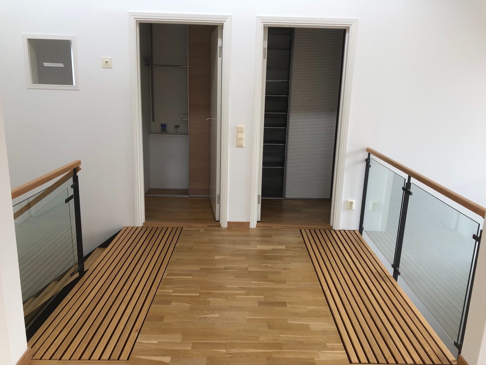 Nowoczesny korytarz z drewnianą podłogą i balustradą ze szkła, prowadzący do dwóch otwartych drzwi z widocznymi wnętrzami garderób z systemami przechowywania.