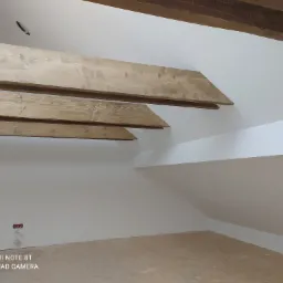 Poddasze po remoncie z widocznymi drewnianymi belkami stropowymi w jasnym odcieniu, białe ściany i przygotowana podłoga przed położeniem wykończenia.