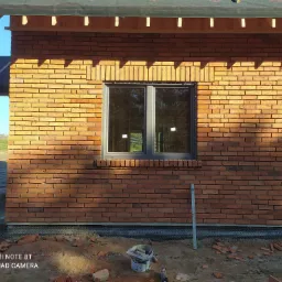 Murowana ściana budynku z oknem, wykonana z cegły klinkierowej w odcieniach czerwieni i pomarańczy, z widocznym poziomym pasem izolacji przy gruncie, oraz narzędziami murarskimi i fragmentami...