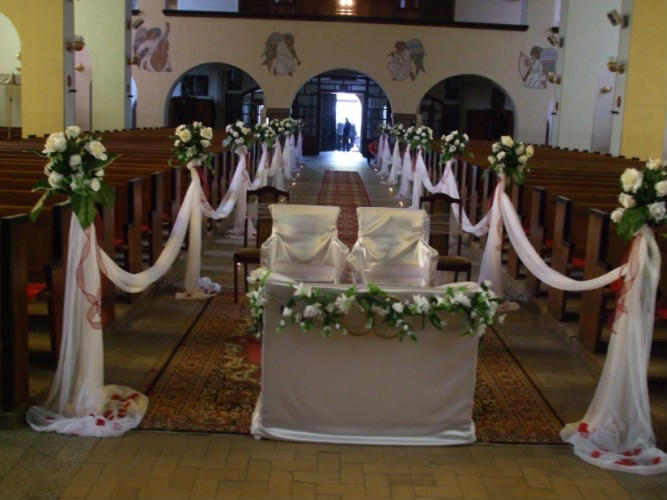 Dekoracja ślubna w kościele: białe kwiaty i tiul zdobią ławki i ołtarz, czerwone akcenty, dywan w orientalny wzór na posadzce.