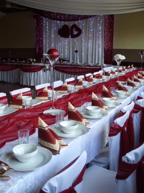 Uroczysta dekoracja sali weselnej w odcieniach bieli i bordo: długi stół z białym obrusem, bordowym bieżnikiem, elegancko złożonymi serwetkami, białymi naczyniami, kieliszkami, krzesła z białymi...