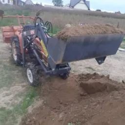Mały traktor z ładowarką czołową, wypełnioną ziemią, w trakcie prac ziemnych na tle wiejskiego krajobrazu z domami.