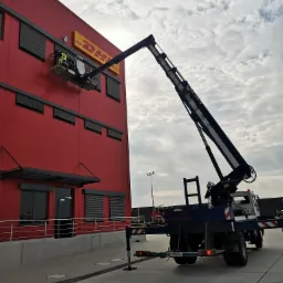 Pracownik w żółtej kamizelce na podnośniku koszowym, czyści lub montuje logo DHL na czerwonym budynku magazynowym z czarnymi żaluzjami.