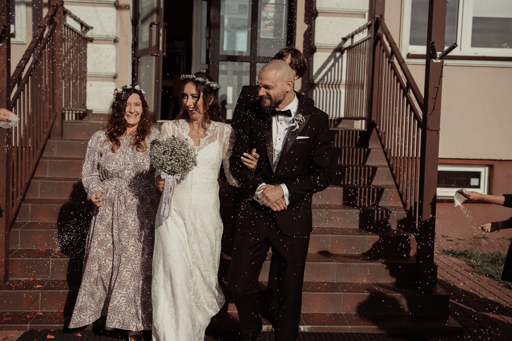 Uśmiechnięta para młoda schodzi po schodach budynku, obsypywana ryżem, w towarzystwie kobiety w kwiecistej sukience i innych osób.