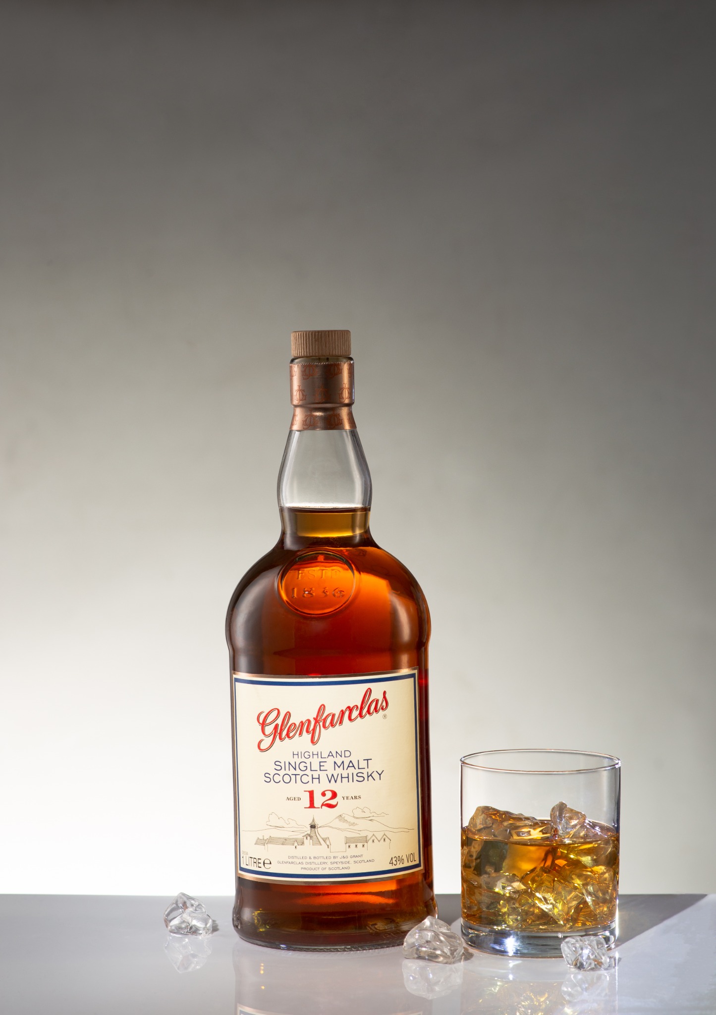 Butelka szkockiej whisky Glenfarclas 12-letniej i szklanka z lodem na błyszczącym blacie, oświetlone studyjnie.