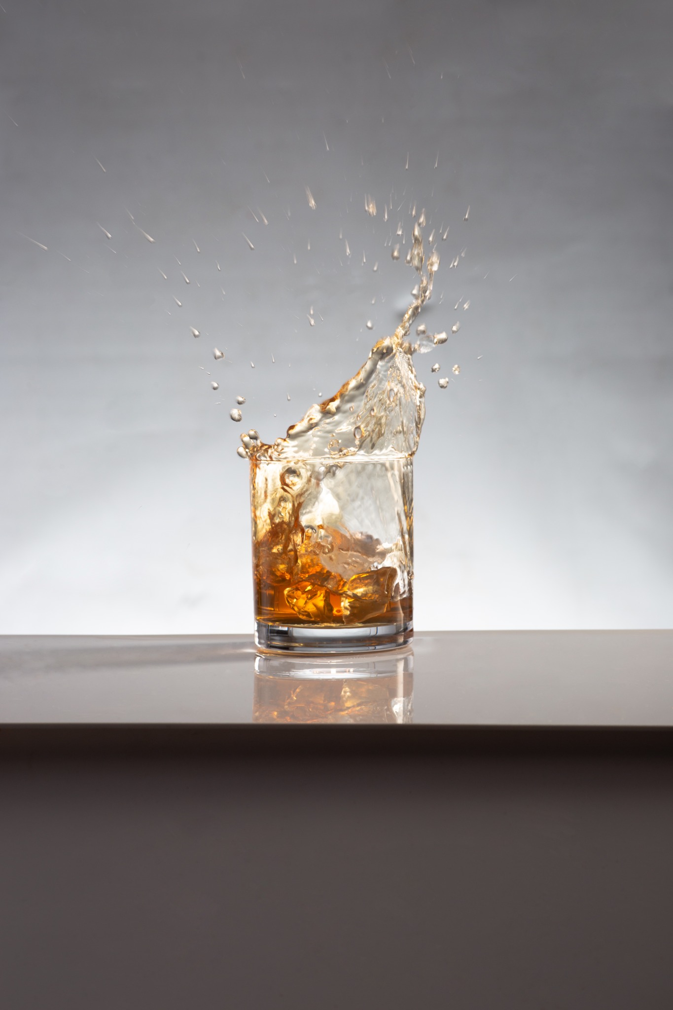 Kieliszek z lodem i brązowym płynem w ruchu, tworzący efektowną fontannę kropli na jasnym tle.