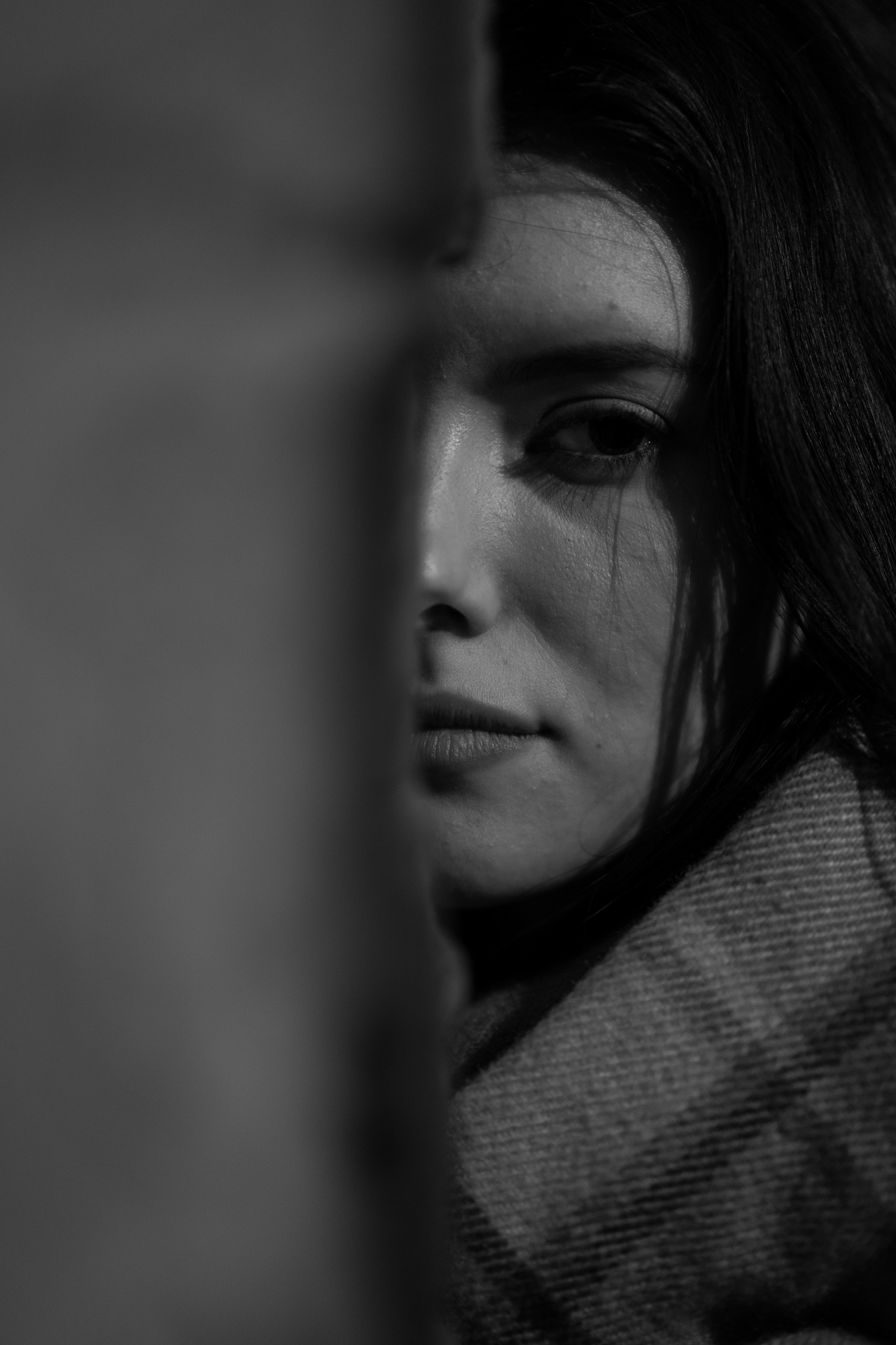 Czarno-białe, portretowe ujęcie kobiety z bliska, częściowo zasłoniętej przez pionowy obiekt, z widoczną fakturą tkaniny na ubraniu.