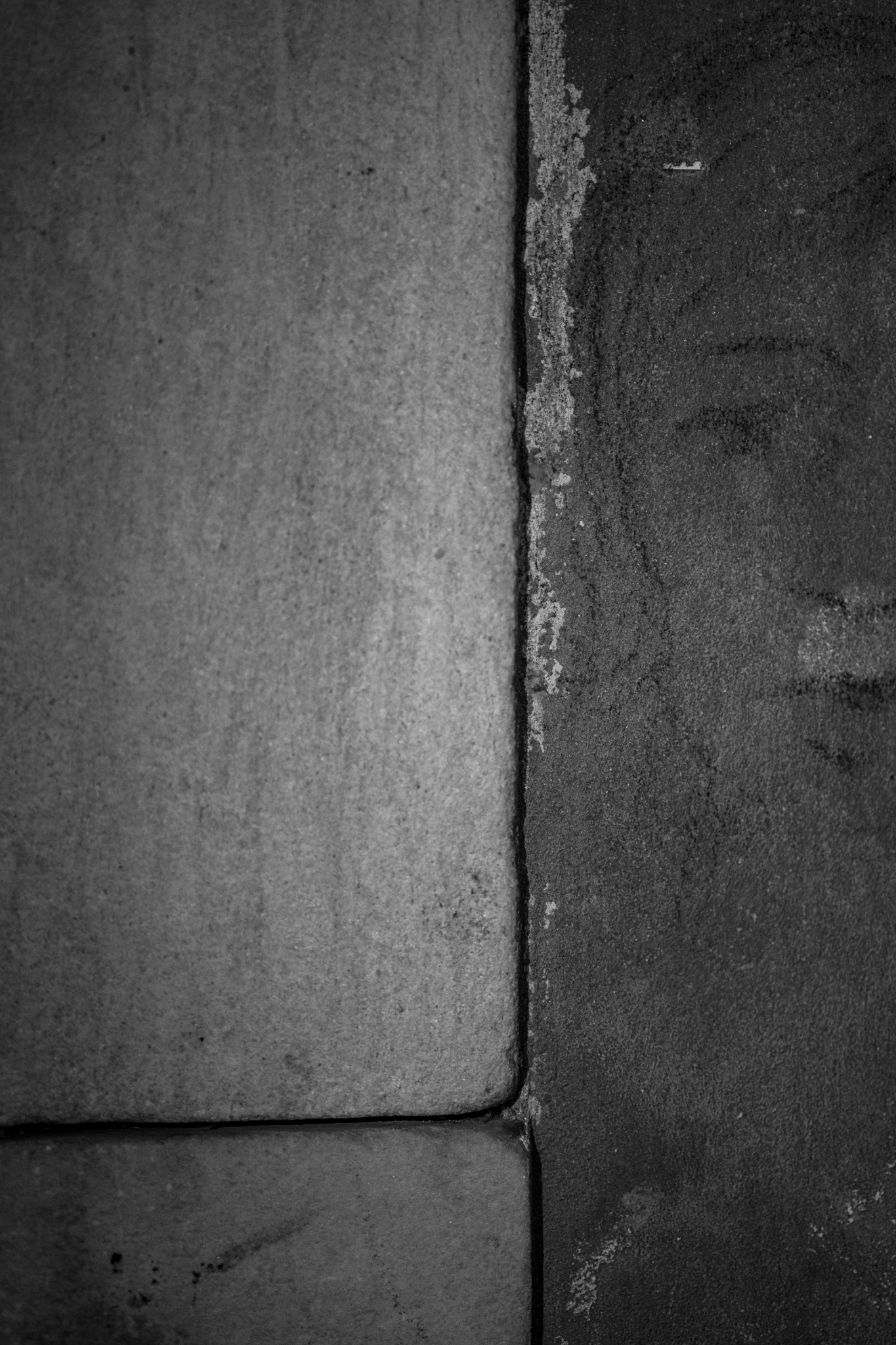 Zbliżenie na połączenie dwóch tekstur: gładkiej, jasnoszarej płyty i chropowatej, ciemnoszarej ściany z odspojoną farbą.