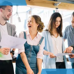 Zespół czterech młodych osób analizuje dokumenty z wykresami w jasnym biurze, kobieta w dżinsowej sukience śmieje się, a mężczyzna w koszuli w kratę wskazuje palcem na arkusz.