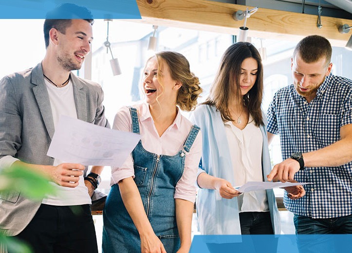 Zespół czterech młodych osób analizuje dokumenty z wykresami w jasnym biurze, kobieta w dżinsowej sukience śmieje się, a mężczyzna w koszuli w kratę wskazuje palcem na arkusz.