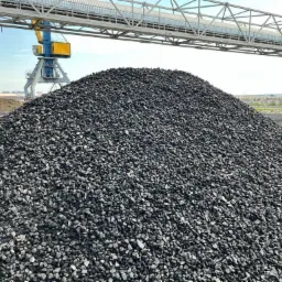 Góra czarnego, drobnego węgla kamiennego na placu składowym, z widocznym żurawiem portowym i estakadą transportową w tle.