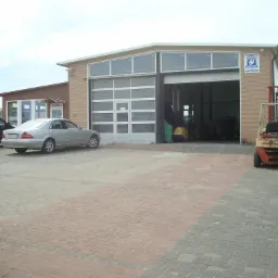 Budynek warsztatu z otwartą bramą, wewnątrz widoczny podnośnik. Na zewnątrz zaparkowane samochody osobowe i wózek widłowy.