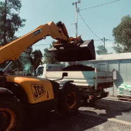 Ż&oacute;łta ładowarka teleskopowa JCB załadowuje ziemię do skrzyni białej ciężar&oacute;wki na tle budynku i palety z workami.