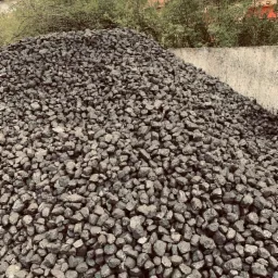 Góra antracytu w kamieniach o nieregularnych kształtach, na tle betonowej ściany i zieleni drzew, częściowo zasłaniająca budynek z czerwonym dachem.