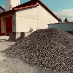 Duży pryzmat ekogroszku na placu składowym, z budynkiem w tle i błękitnym niebem.