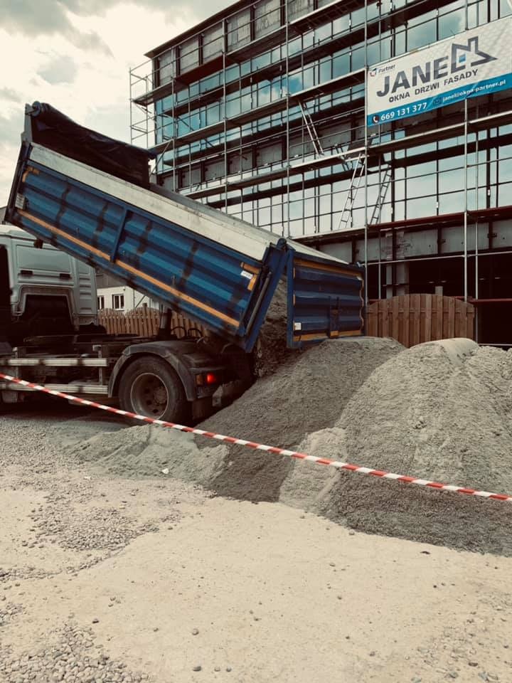 Wyładowywany z wywrotki materiał sypki, prawdopodobnie piasek lub żwir, na placu budowy z rusztowaniami i budynkiem w tle.