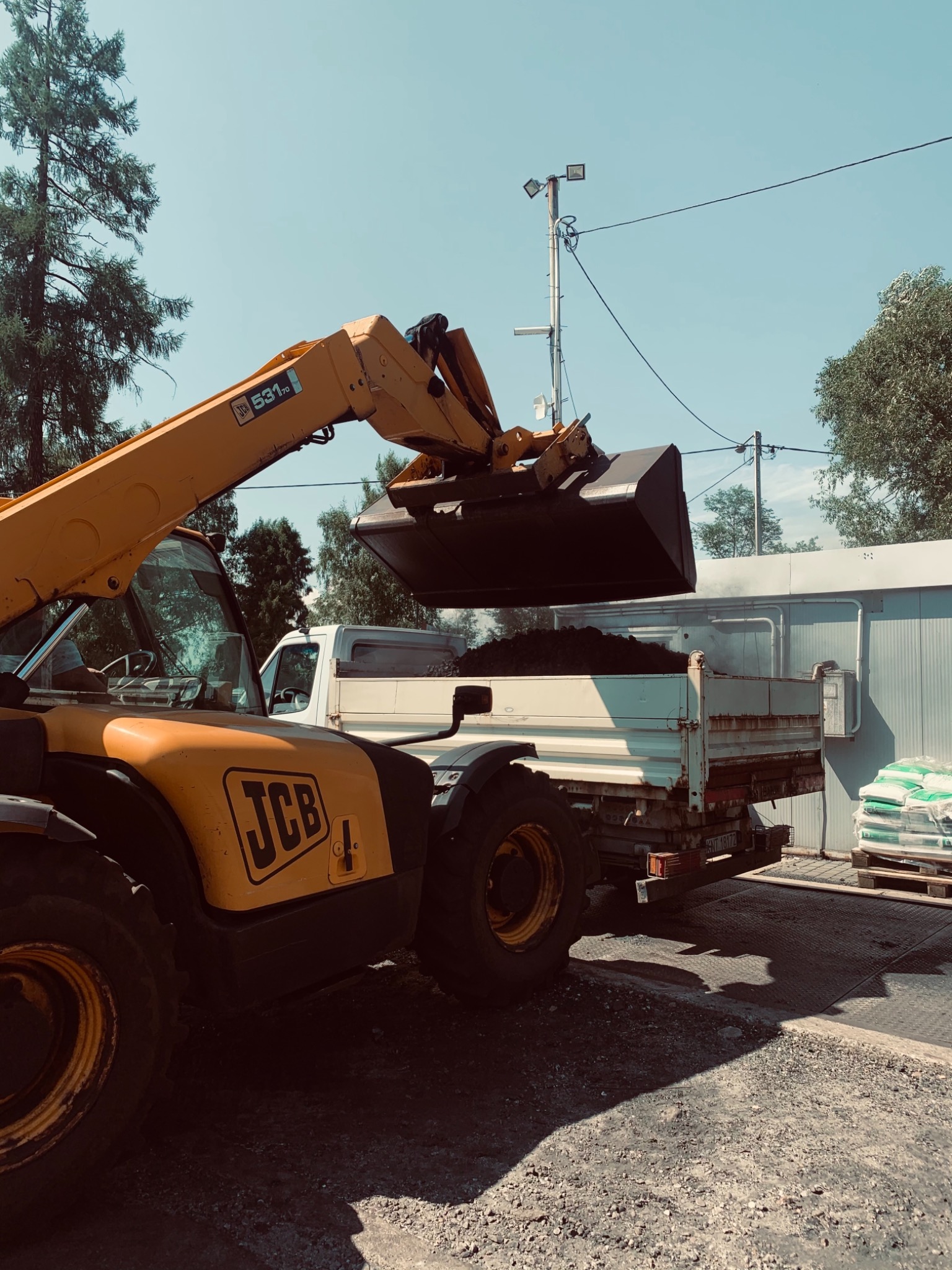 Żółta ładowarka teleskopowa JCB załadowuje ziemię do skrzyni białej ciężarówki na tle budynku i palety z workami.