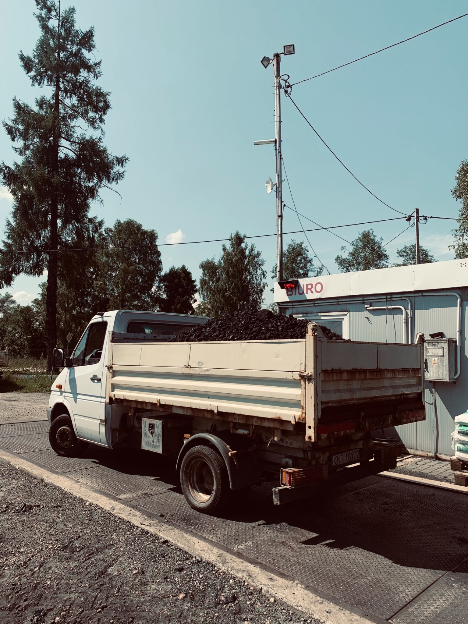 Biały samochód dostawczy z otwartą skrzynią załadowaną czarnym węglem, stojący na wadze samochodowej przy budynku biurowym w słoneczny dzień.