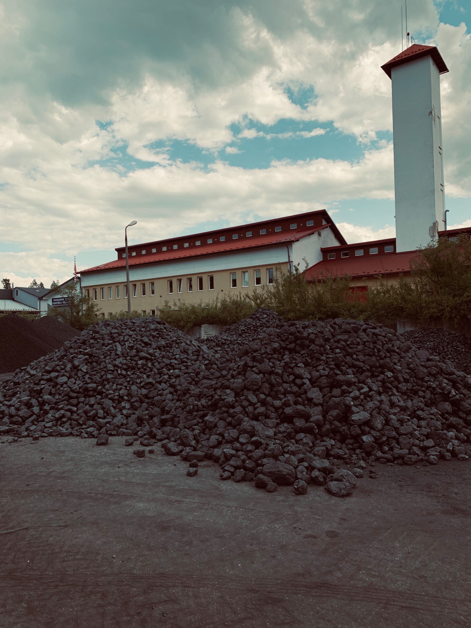 Dwa pryzmy czarnego węgla na placu składowym, w tle budynek z wieżą i pochmurne niebo.