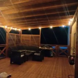 Zadaszony taras z drewnianą podłogą i konstrukcją, oświetlony girlandą żarówek, z meblami ogrodowymi i ażurową ścianką, w tle widoczny basen.