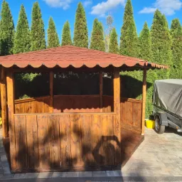 Drewniana altana z czerwoną dachówką na posesji, w tle wysokie tuje i przyczepa z plandeką.