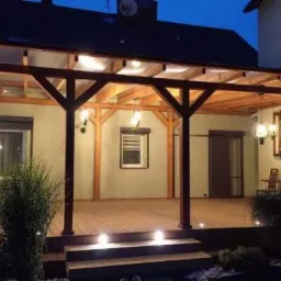 Oświetlona drewniana pergola z przezroczystym zadaszeniem i drewnianym tarasem, widok wieczorny z lampami wiszącymi i punktowymi, dekoracyjna roślinność wzdłuż obrzeży.