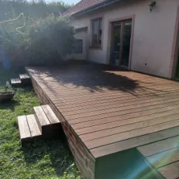 Rozległy, ciemnobrązowy taras z desek, z trzema stopniami prowadzącymi na trawnik, przylegający do jasnego domu z brązowymi oknami i drzwiami przesuwnymi.