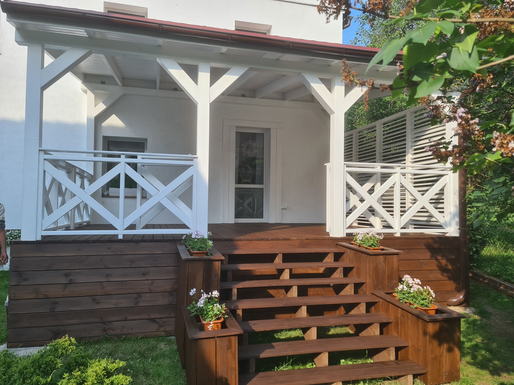 Zadaszony taras drewniany z białymi elementami konstrukcyjnymi, schodami z donicami z kwiatami i drewnianą balustradą z kratownicą.