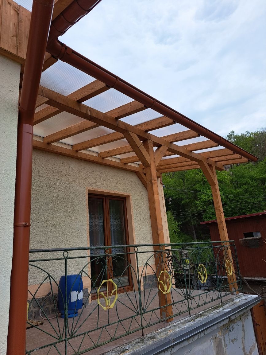 Drewniana konstrukcja z przezroczystym zadaszeniem nad balkonem z metalową balustradą z dekoracyjnymi elementami, widok z dołu na tle zieleni.