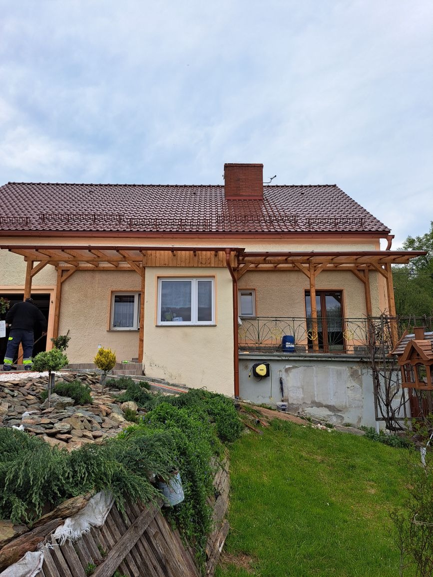 Dom z beżową elewacją i drewnianą konstrukcją wspierającą dach, widoczny z boku, z zielonym ogrodem skalnym na pierwszym planie.