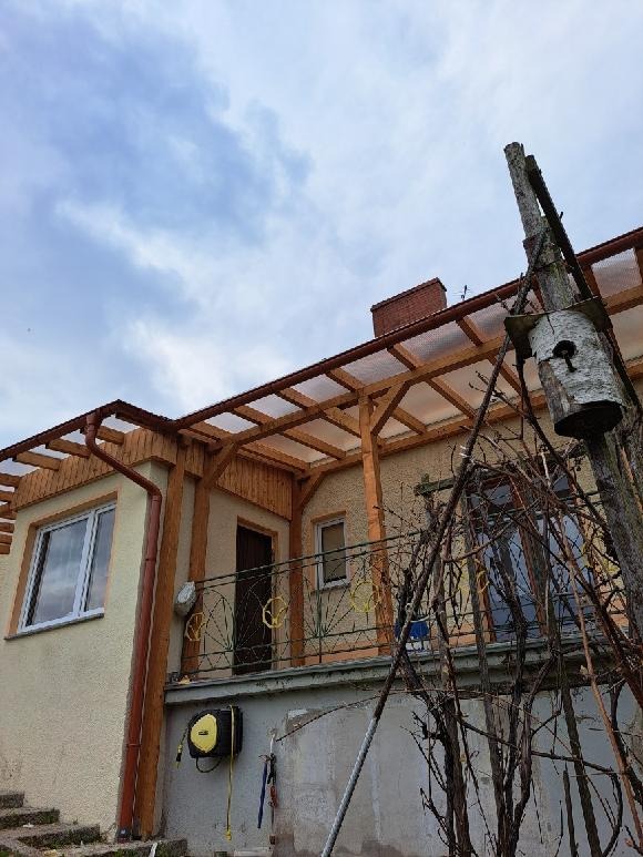 Drewniana konstrukcja z przezroczystym zadaszeniem przy balkonie domu jednorodzinnego, widoczna skrzynka lęgowa dla ptaków na słupie, zwinięty wąż ogrodowy na ścianie.