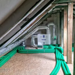 Instalacja rekuperacji Vent-Axia na poddaszu z widocznymi zielonymi przewodami wentylacyjnymi i aluminiowymi rurami, surowa konstrukcja dachu z izolacją.
