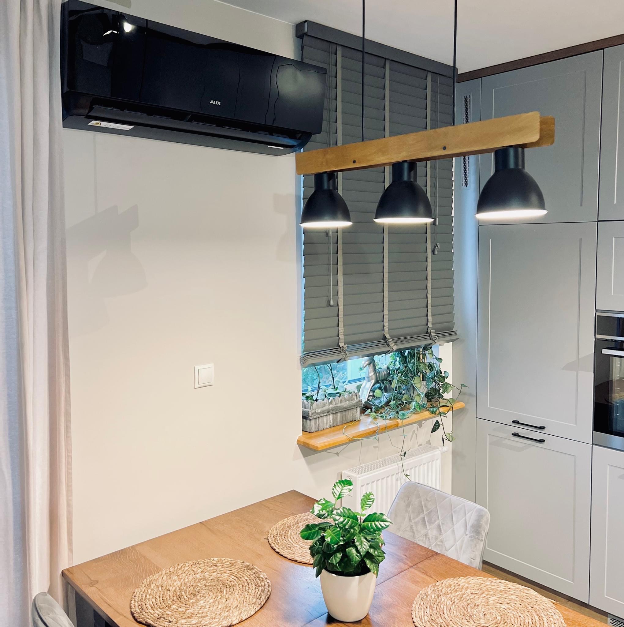 Czarna jednostka klimatyzacyjna AUX zamontowana w nowoczesnym wnętrzu nad drewnianym stołem z plecionymi podkładkami, oświetlona trzema wiszącymi lampami, szare żaluzje w oknie z roślinami i szare...