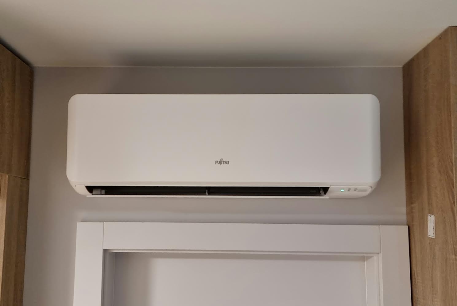 Biała jednostka klimatyzacji Fujitsu zamontowana nad futryną drzwi w pomieszczeniu o szarych ścianach i drewnianych elementach wykończeniowych.