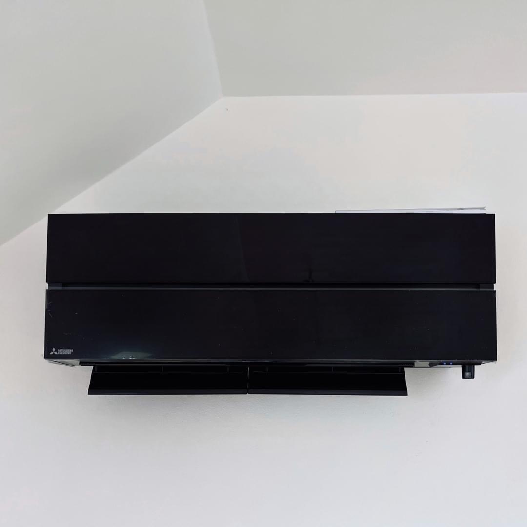 Czarny klimatyzator Mitsubishi zawieszony na białej ścianie w rogu pomieszczenia, widoczne otwarte żaluzje.