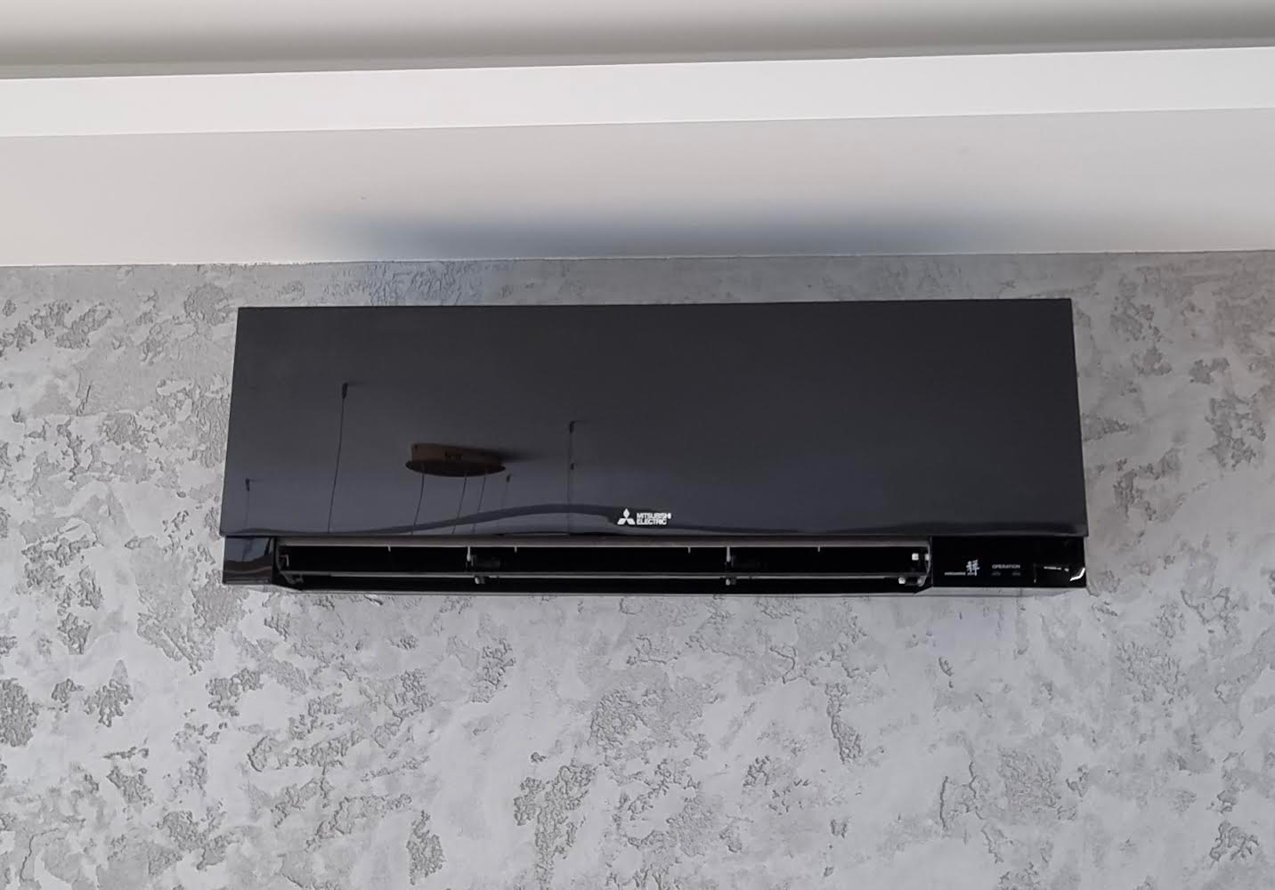 Czarny klimatyzator Mitsubishi Electric zamontowany na szarej ścianie o teksturze betonu, widoczny pod sufitem.