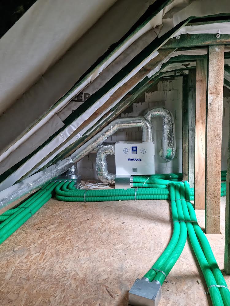 Instalacja rekuperacji Vent-Axia na poddaszu z widocznymi zielonymi przewodami wentylacyjnymi i aluminiowymi rurami, surowa konstrukcja dachu z izolacją.