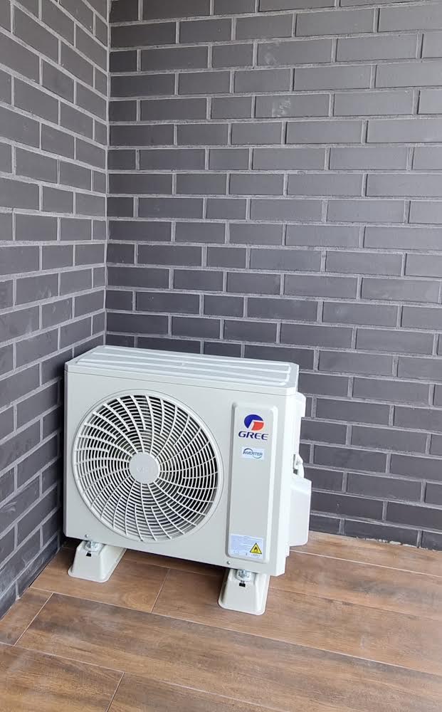 Biała jednostka zewnętrzna klimatyzatora marki Gree, zamontowana na drewnopodobnej podłodze przy szarej, ceglanej ścianie.