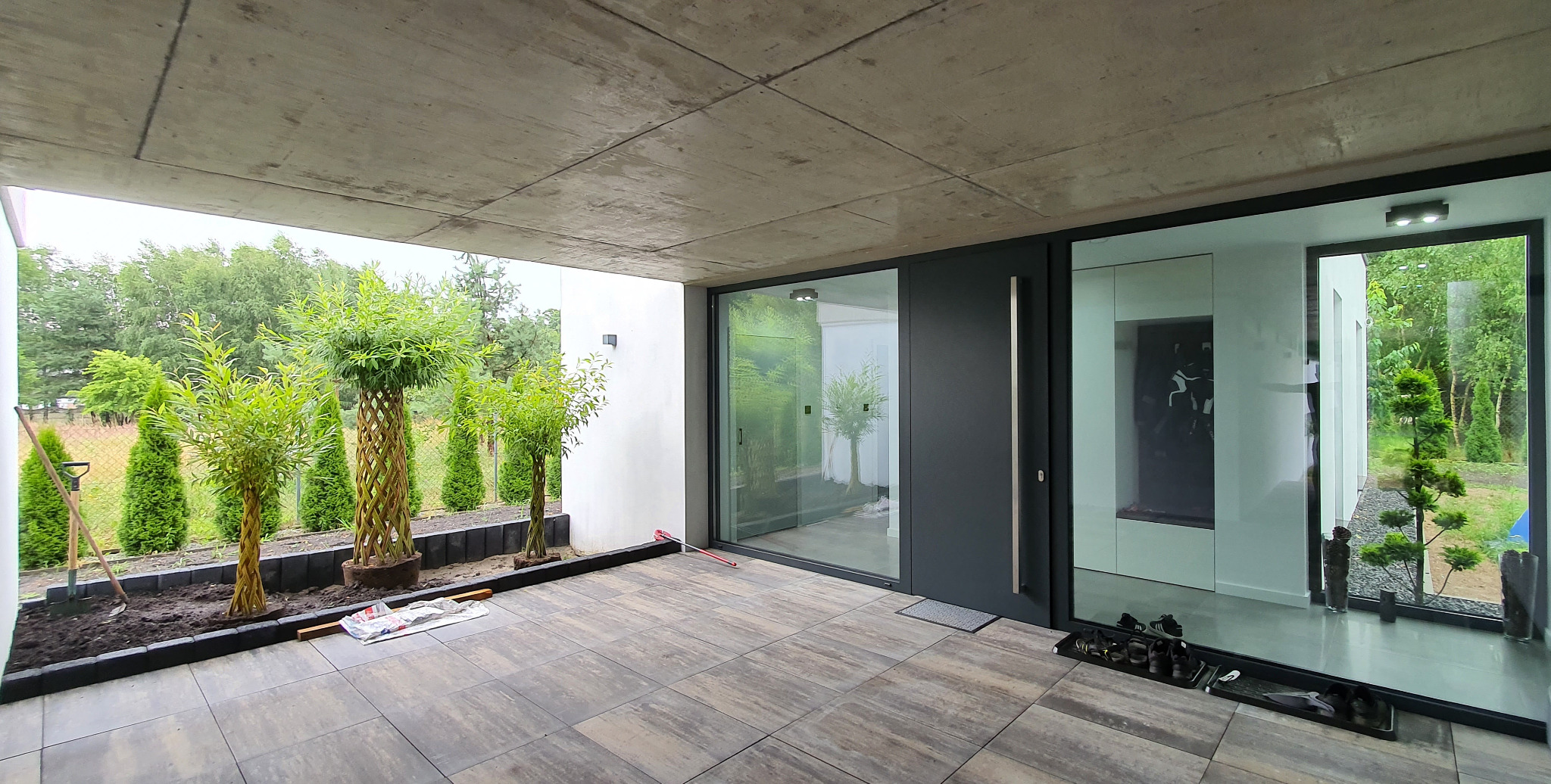 Nowoczesny taras z szarą płytką, minimalistyczne wejście do domu z dużymi przeszkleniami, drzewka formowane w plecionki, narzędzia ogrodnicze obok przygotowanego miejsca do sadzenia.