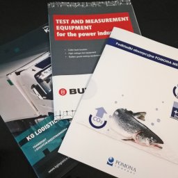 Powielarnia MADAS - Stos ulotek i broszur firmowych, w tym ulotka firmy transportowej KG Logistics, broszura z wyposażeniem pomiarowym i ulotka o podkładkach absorpcyjnych Pomona Mega CO2 z grafiką ryby.