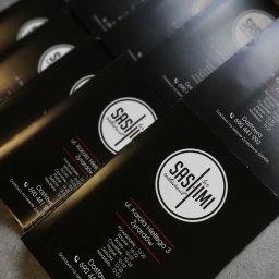 Powielarnia MADAS - Stos czarnych wizytówek restauracji Sashimi z białym logo i informacjami o godzinach otwarcia oraz dostawie na terenie Żyrardowa, ułożonych pod kątem na szarym tle.